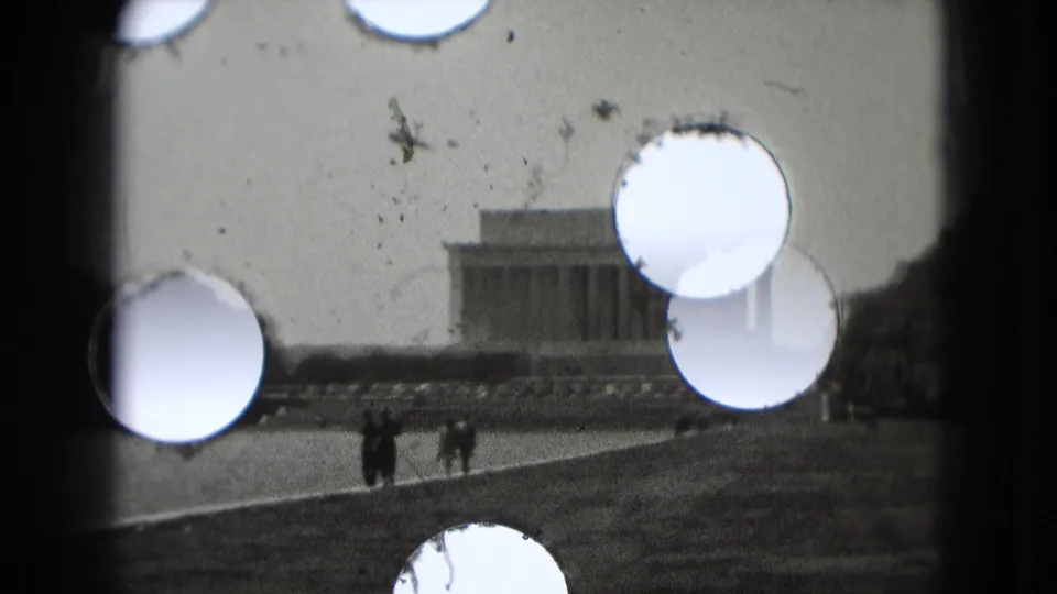 The Lincoln Memorial appears in the distance in Washington DC, with visitors walking in the foreground and bright film perforations crossing the 8mm frame.