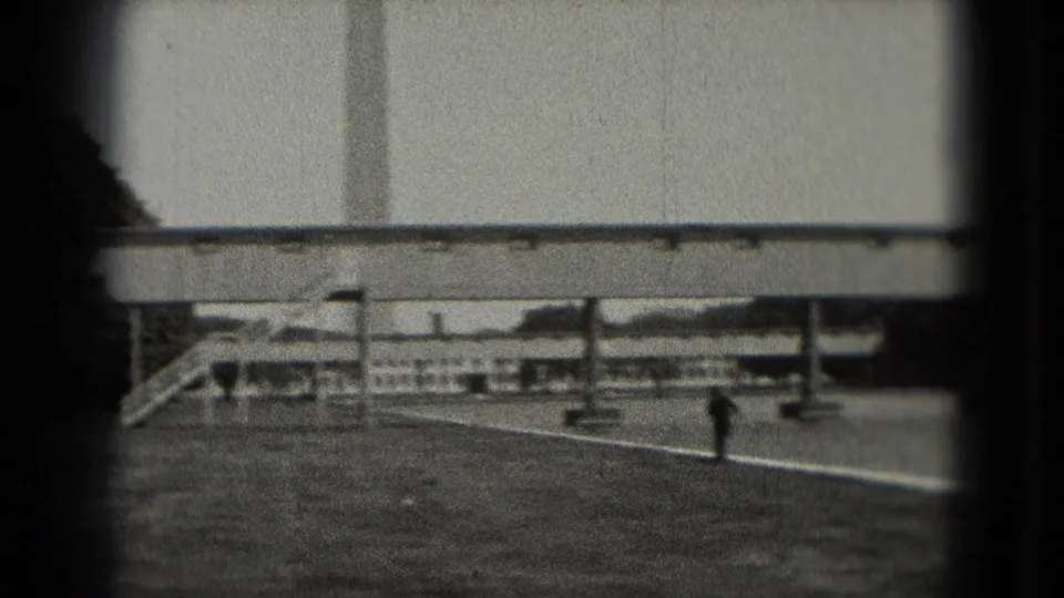 Black-and-white 1975 Washington, DC home-movie frame with a distant man crossing a paved riverside or plaza area below a long elevated structure.