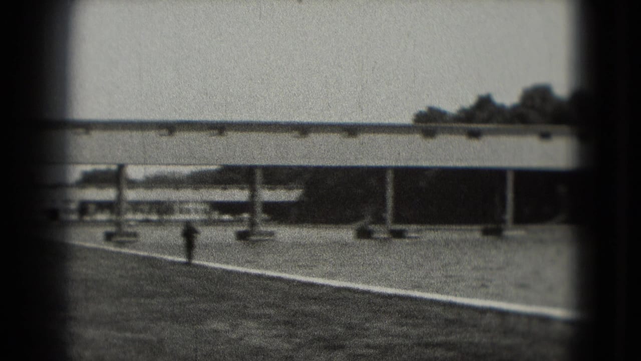 Black-and-white Washington, DC frame showing a quiet paved waterside area, railings, columns, and a long bridge or walkway in the background.