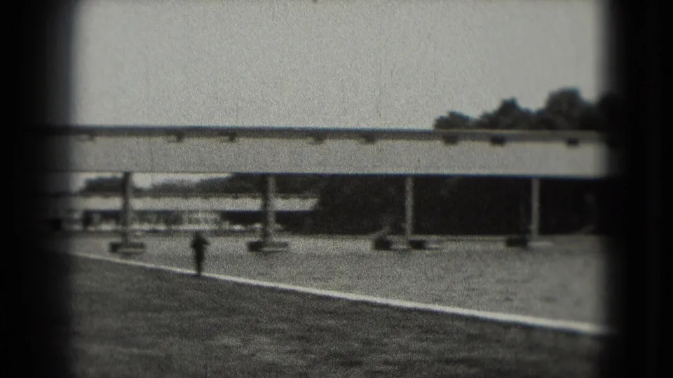 Black-and-white Washington, DC frame showing a quiet paved waterside area, railings, columns, and a long bridge or walkway in the background.