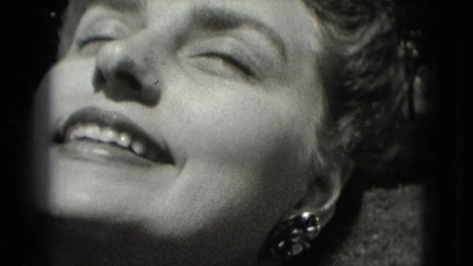 1975 Washington, DC home-movie frame of an adult woman smiling with eyes closed, decorative earring visible, and strong sunlight across her face.