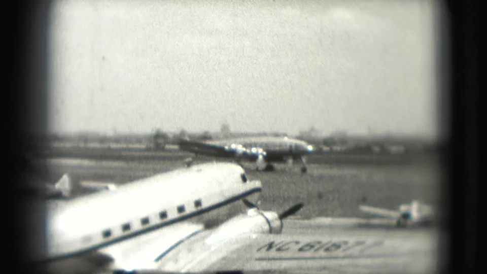 1975 airport tarmac footage shows a propeller aircraft in the foreground and additional planes farther back, filmed from a distant home-movie viewpoint.