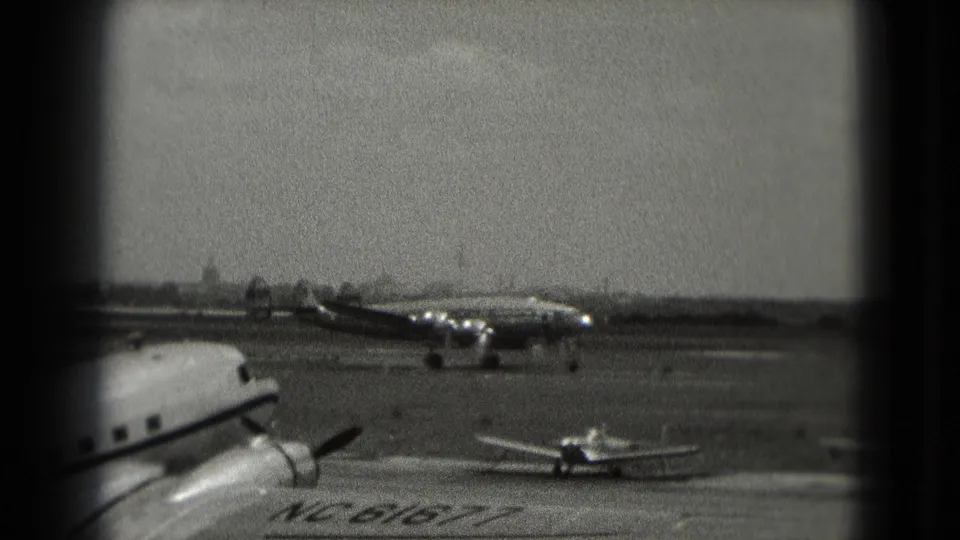 1975 Washington DC 8mm frame showing a propeller airliner taxiing across an airfield with smaller aircraft in the foreground.