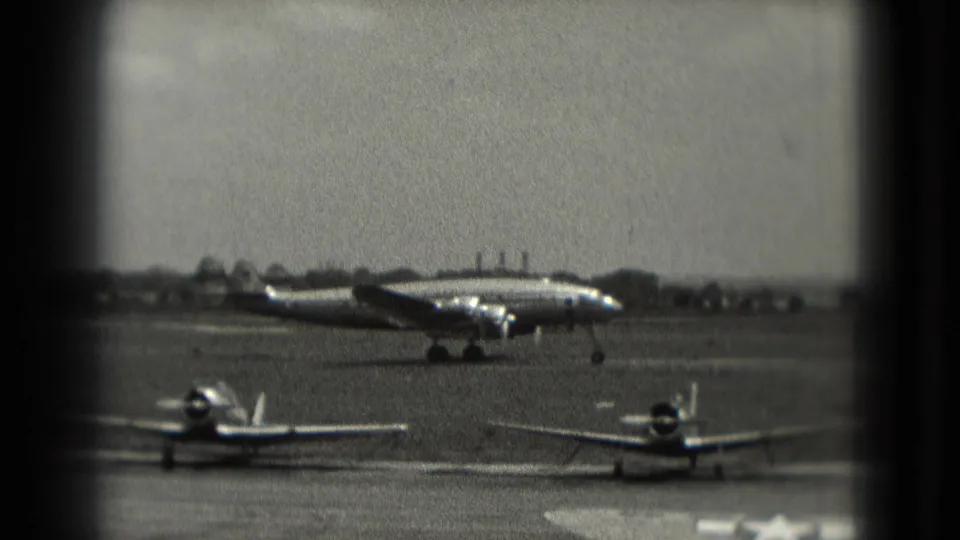 Large propeller aircraft crosses the Washington, DC airfield runway in a grainy 1975 8mm frame with small planes parked nearby.