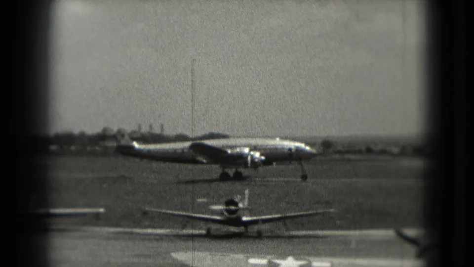 1975 Washington, DC home-movie frame with a propeller aircraft taxiing behind a small foreground aircraft on an open runway.