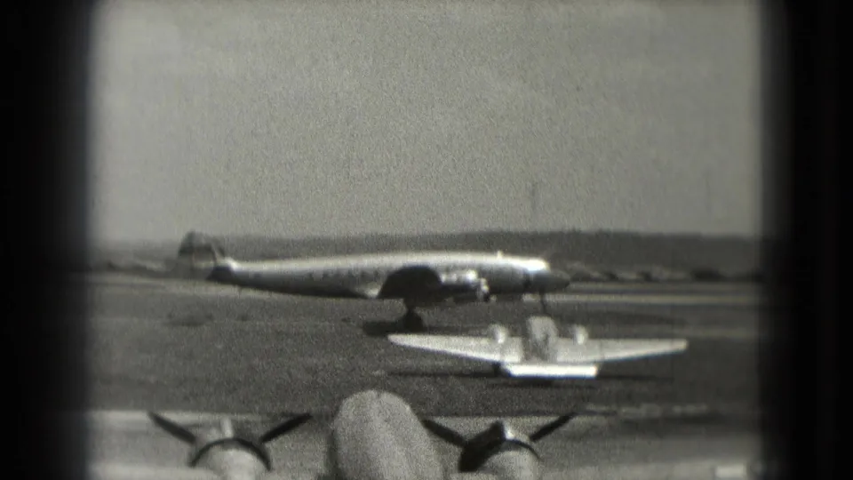 1975 Washington, DC airfield view with a large propeller aircraft, a smaller plane, and foreground aircraft engines in soft 8mm focus.
