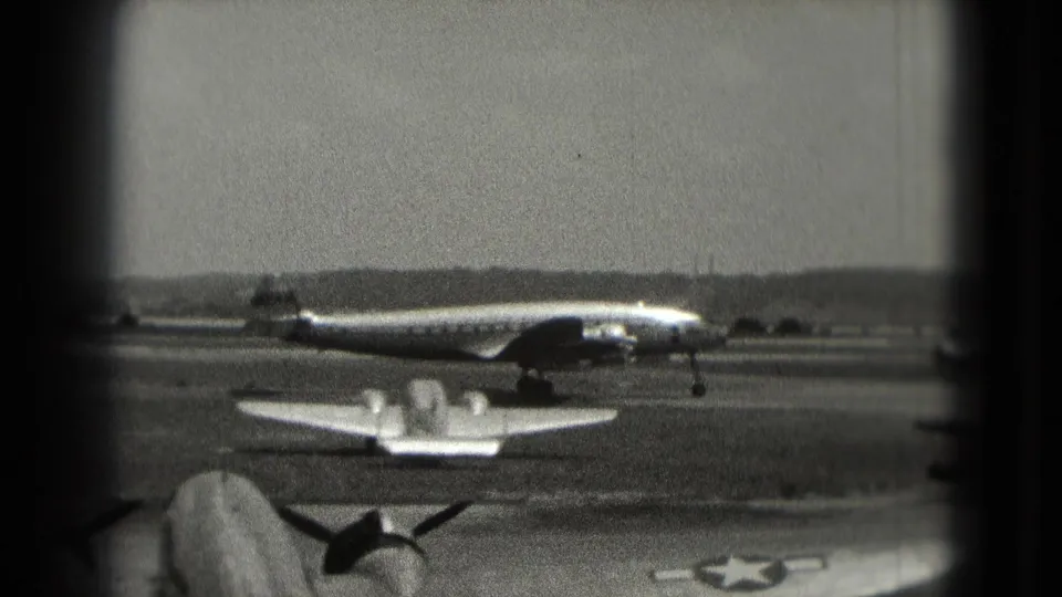 1975 Washington, DC runway scene showing a propeller aircraft passing behind a smaller plane, seen through grainy 8mm framing.