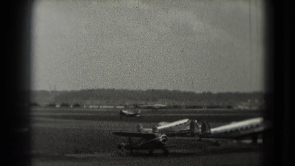 1975 Washington-area 8mm frame showing a propeller plane on the tarmac, a mobile stairway, distant airfield, and hazy sky.