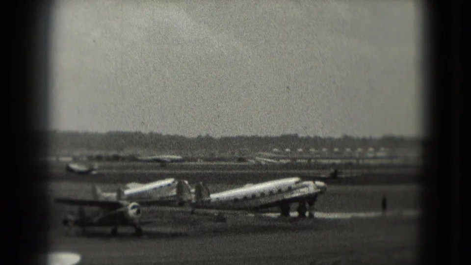 Soft 8mm Washington-area airport frame showing a propeller plane, mobile stairs, open tarmac, and small distant figures.