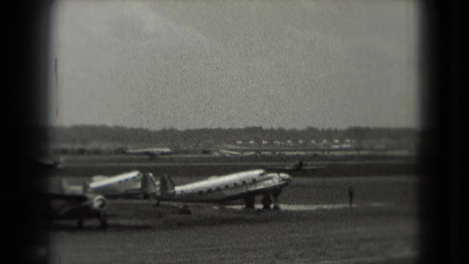 1975 Washington-area 8mm frame with a parked propeller plane, mobile stairway, distant airfield traffic, and grainy sky.