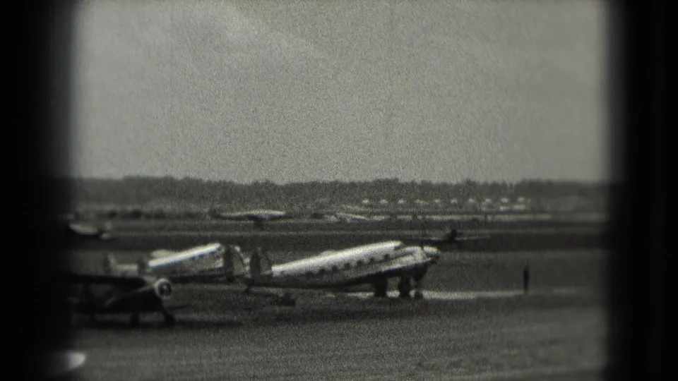 1975 8mm frame from Washington, DC area footage showing a vintage twin-engine propeller aircraft on an airfield ramp with distant figures.