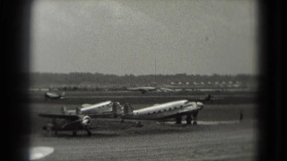 Vintage propeller aircraft appears parked on a Washington, DC area airfield ramp in grainy 1975 8mm footage, with distant ground activity.