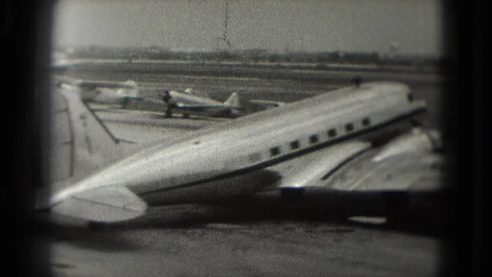 Black-and-white 8mm frame from 1975 showing a small passenger plane fuselage in the foreground and parked aircraft behind it on a Washington, DC tarmac.