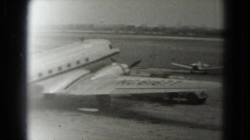 Soft black-and-white 8mm view of a parked passenger aircraft on a Washington, DC airport tarmac in 1975, with another small plane visible in the distance.