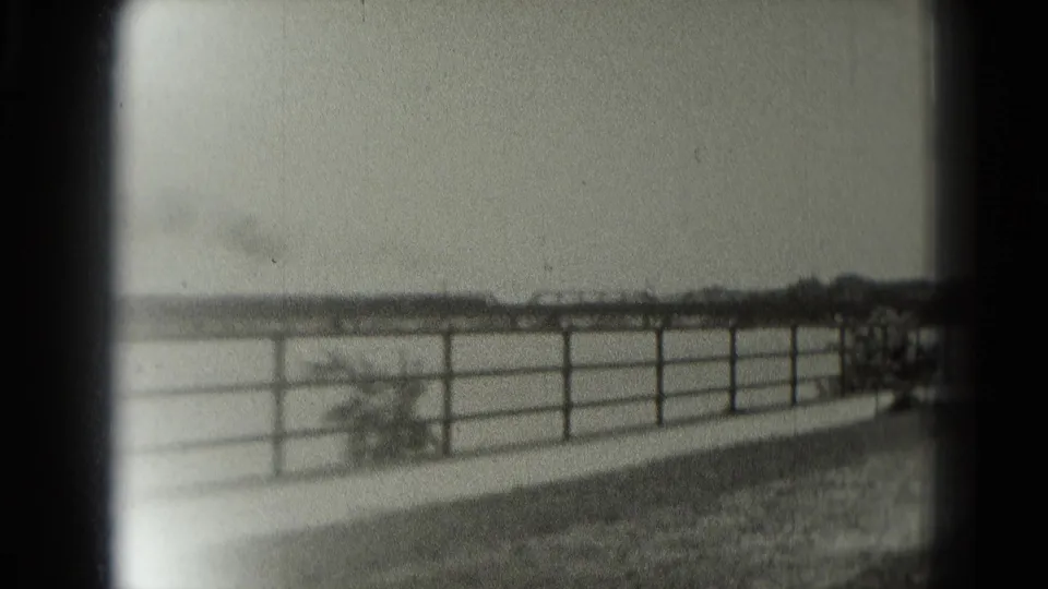 A metal railing borders grass beside open water in Washington DC, seen through soft 8mm black-and-white home-movie footage from 1975.