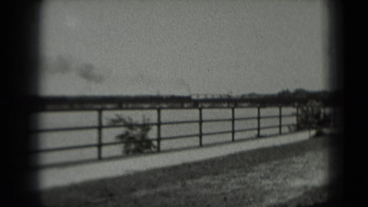 Metal fence beside open water or field with a distant smoky rail line on the horizon.