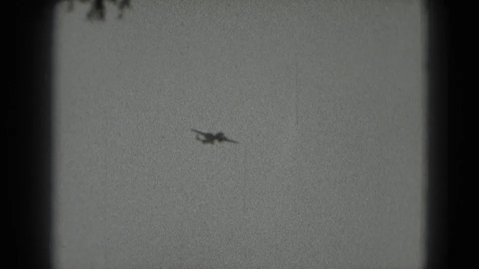 A small airplane appears as a dark silhouette against an overcast sky in this 1975 8mm home-movie frame.
