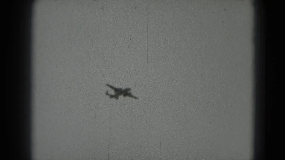 The aircraft remains visible in black-and-white 8mm footage, silhouetted against a plain overcast sky.
