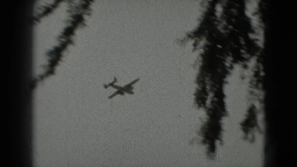 The plane remains visible in the pale sky with dark tree silhouettes and film-edge shadows in the 1975 8mm footage.