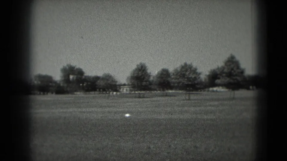 1975 Washington, DC 8mm frame showing an open grassy field, distant tree line and a small pale object in the foreground.