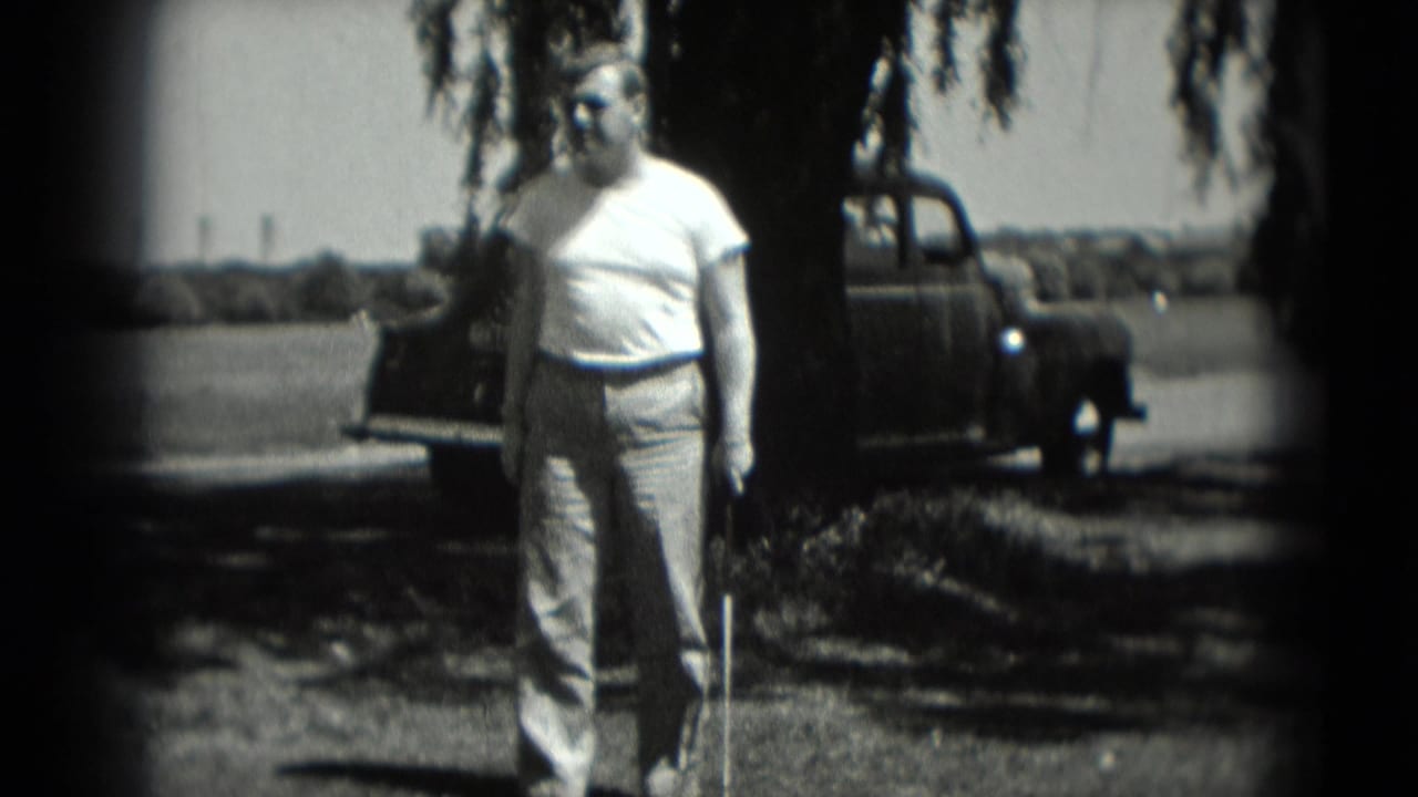 Man holding a golf club near a large tree with a pickup truck behind him.