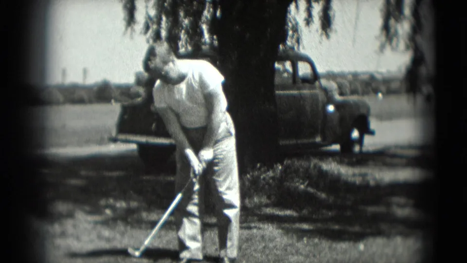 1975 Washington, DC home-movie frame showing a man addressing a golf ball near a tree and parked pickup truck.