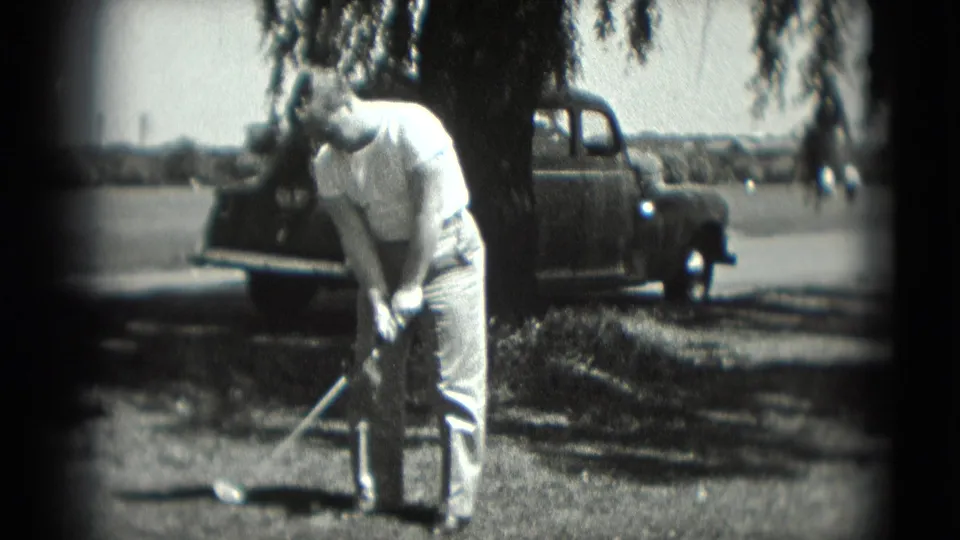 1975 Washington, DC home-movie frame of a man swinging a golf club beside a tree with a parked pickup truck behind him.
