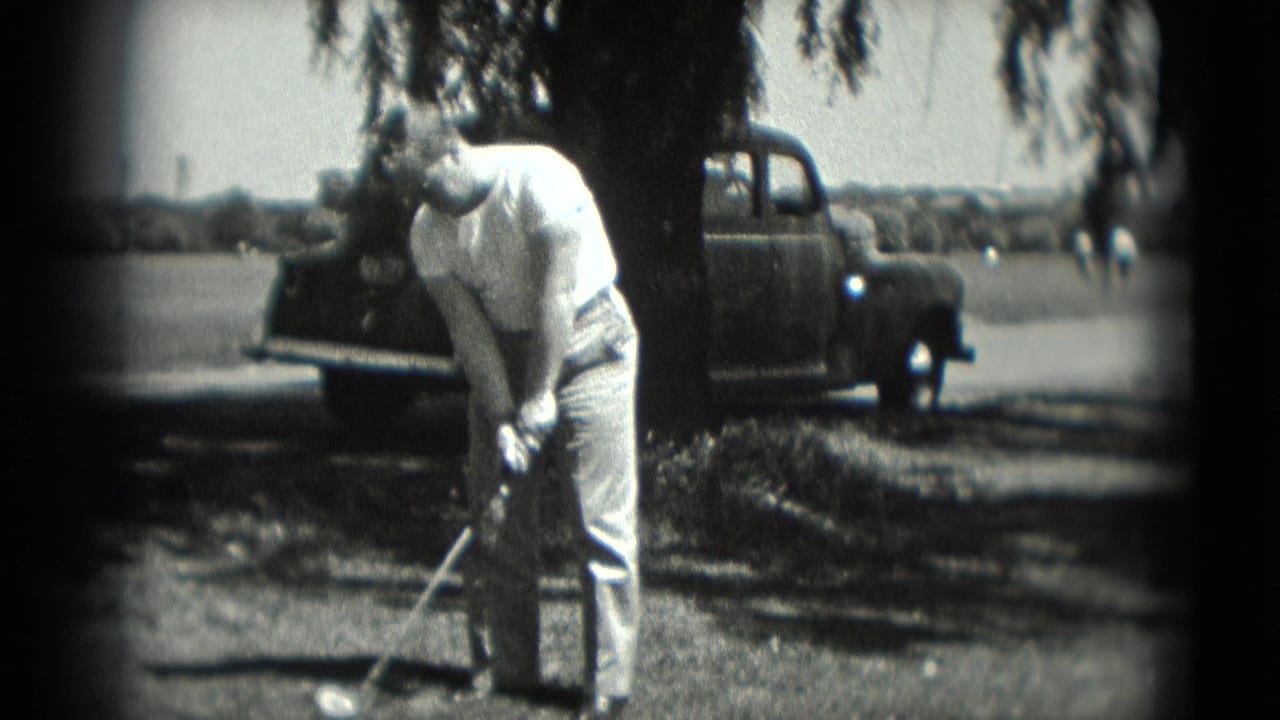 In Washington, DC 1975 home-movie footage, an adult man prepares a golf swing near a parked older car and leafy tree shade.