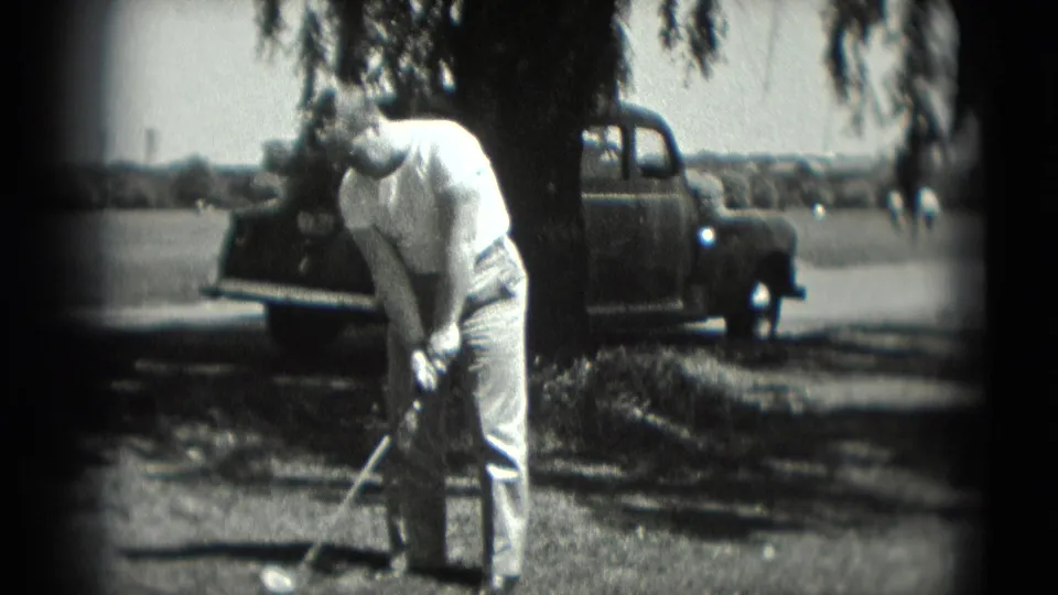 In Washington, DC 1975 home-movie footage, an adult man prepares a golf swing near a parked older car and leafy tree shade.