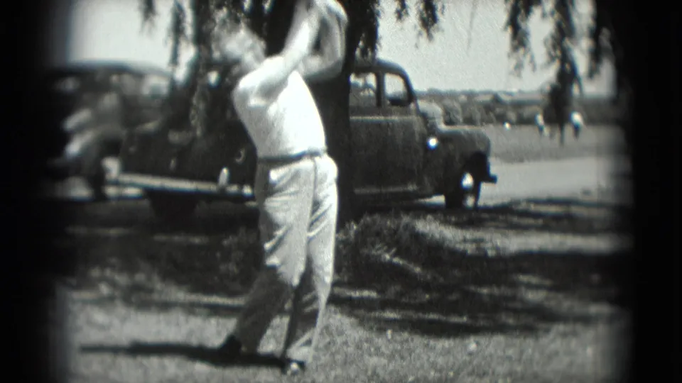 An adult man swings a golf club in the 1975 Washington, DC home-movie scene, with an older car and roadside background visible.
