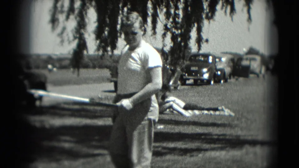 The man holds a golf club in a 1975 Washington, DC recreation scene, with a sunbather on a blanket and parked older cars behind him.