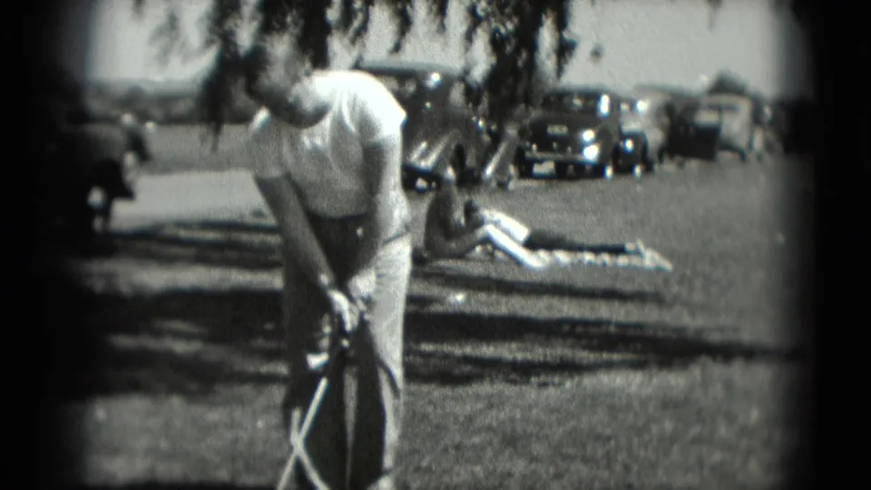 A man bends into position with a golf club in the Washington, DC 1975 home movie, while a person lies on the grass near parked cars.