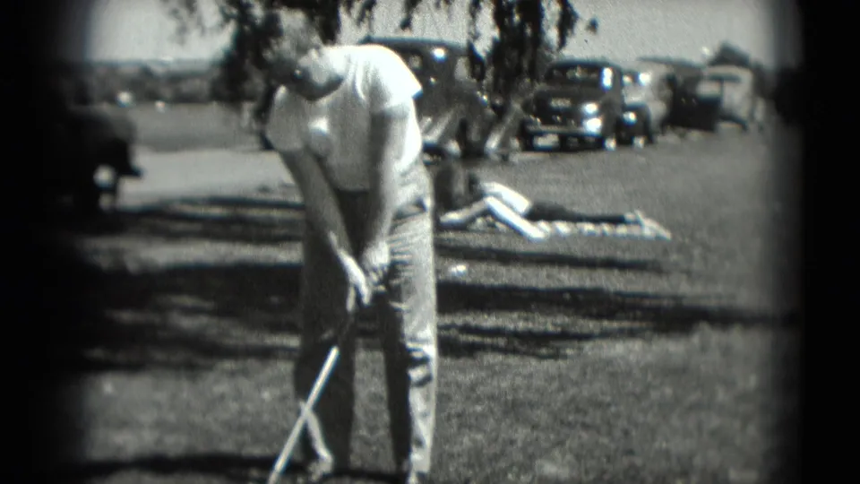 The man sets up for a golf swing on a grassy Washington, DC recreation area in 1975, with a sunbather and older parked cars visible behind him.