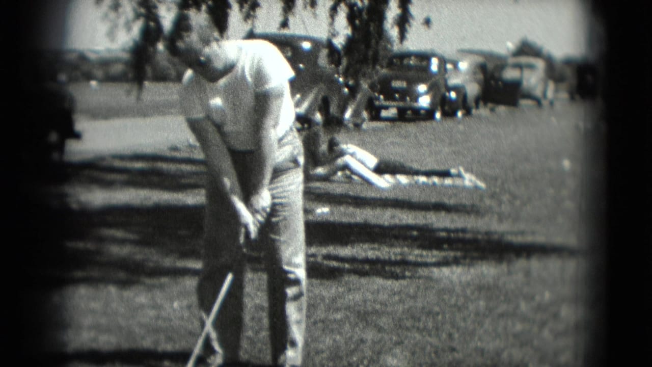 Man in a white shirt bends over a golf club on grass with parked cars behind him.