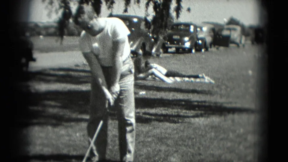 In 1975 Washington, DC home-movie footage, a man addresses a golf ball on grass while cars and a person on a blanket sit in the background.