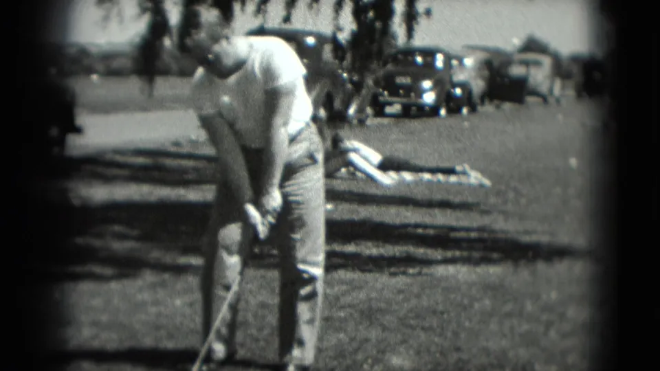 The golfer prepares his swing in a Washington, DC outdoor scene, with mid-1970s cars and a person lying on a blanket behind him.