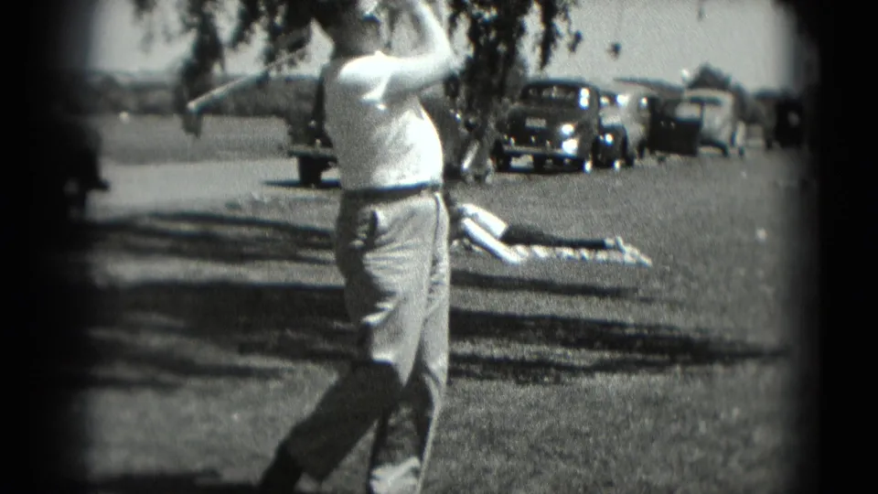 In the 1975 8mm scene, the man swings through with his golf club while a person on a blanket and parked cars remain in the background.
