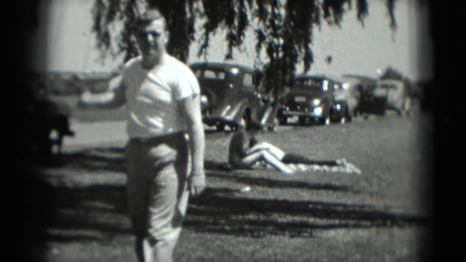 The golfer stands after his swing in the Washington, DC home movie, with tree shade, parked cars, and a person on a blanket visible behind him.