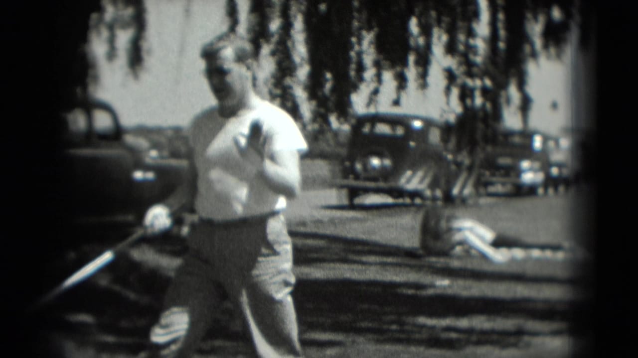 Man holding a golf club on grass with parked cars and hanging tree branches behind him.