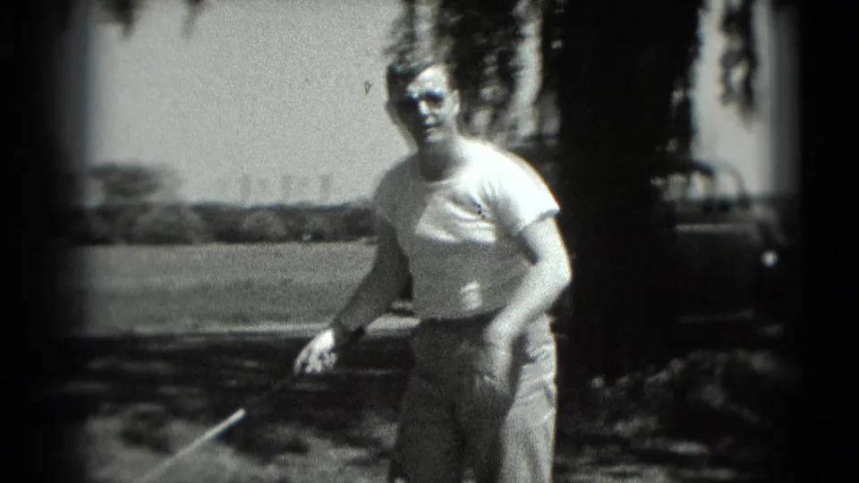 1975 Washington DC 8mm frame of a man holding a golf club near shaded tree cover, with open grass and a parked car visible behind him.