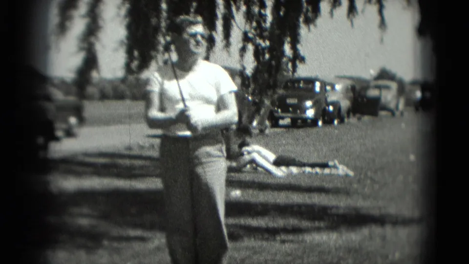 1975 Washington DC 8mm frame showing a man with a golf club facing the camera, parked cars behind him, and another person reclining on the grass.