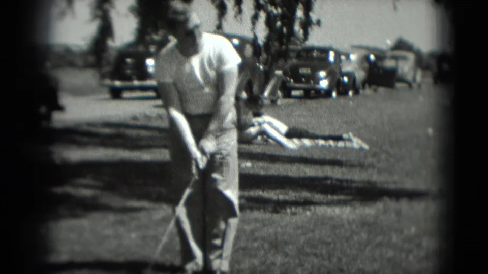 1975 Washington DC 8mm frame of a man leaning into a golf stance, with parked cars and a person lying on the grass in the background.