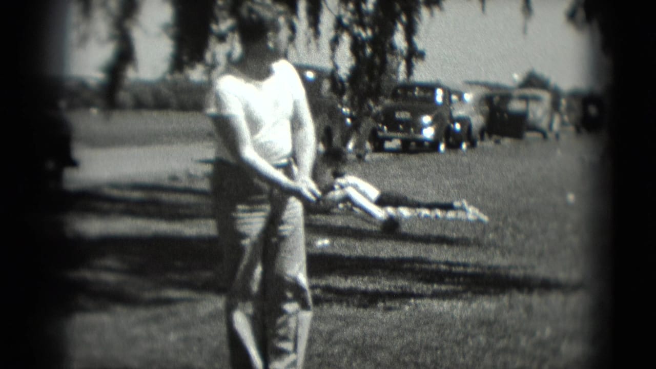 Man in a white shirt lines up a golf swing on grass near parked cars.