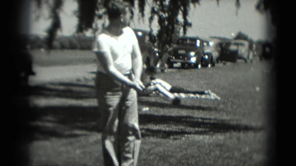 In supported 1975 Washington, DC footage, a man prepares a golf swing on grass while parked cars and a reclining person appear behind him.