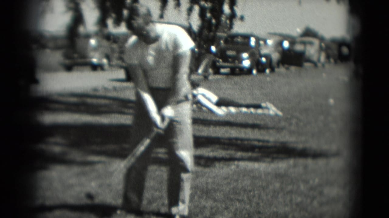 The man stands over his golf club in the 1975 Washington, DC scene, with a person on a blanket and vintage cars in the background.