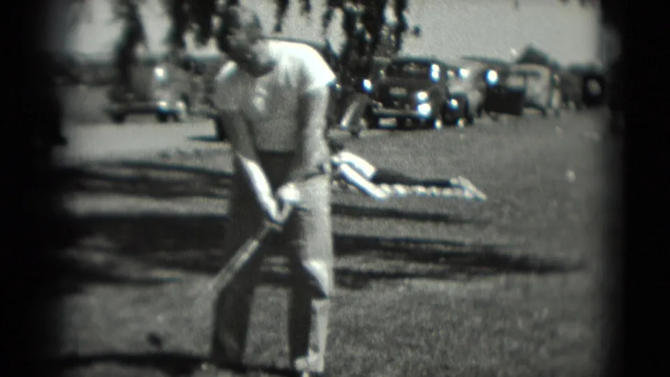 The man stands over his golf club in the 1975 Washington, DC scene, with a person on a blanket and vintage cars in the background.