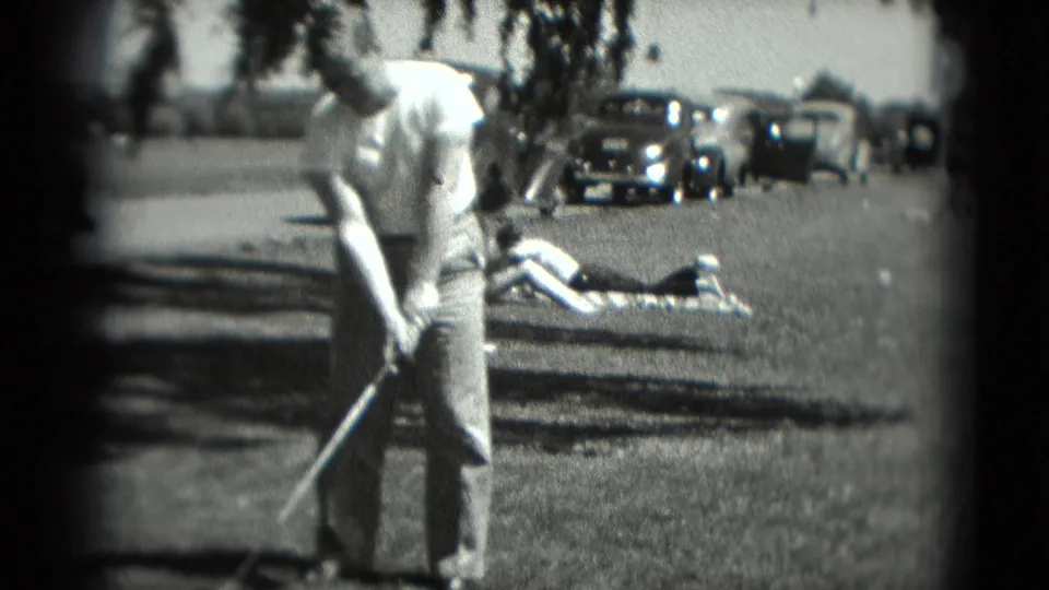 A man holding a golf club is framed close in the foreground, with a reclining person, trees, and parked cars behind him in Washington, DC.