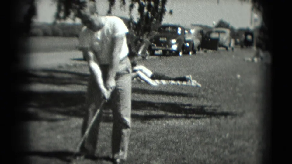 In 1975 Washington, DC home-movie footage, a man stands with a golf club on grass as parked automobiles and a reclining person appear behind him.