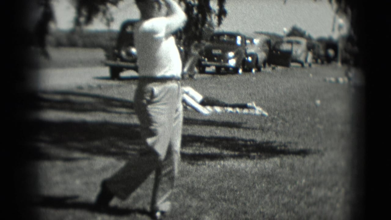 Man follows through with a golf swing on grass, with cars and a person on a blanket in the background.