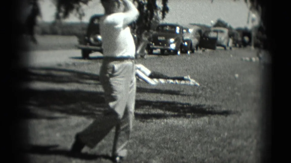 A man in light trousers completes a golf swing in 1975 Washington, DC footage, with vintage cars and a resting person visible behind him.